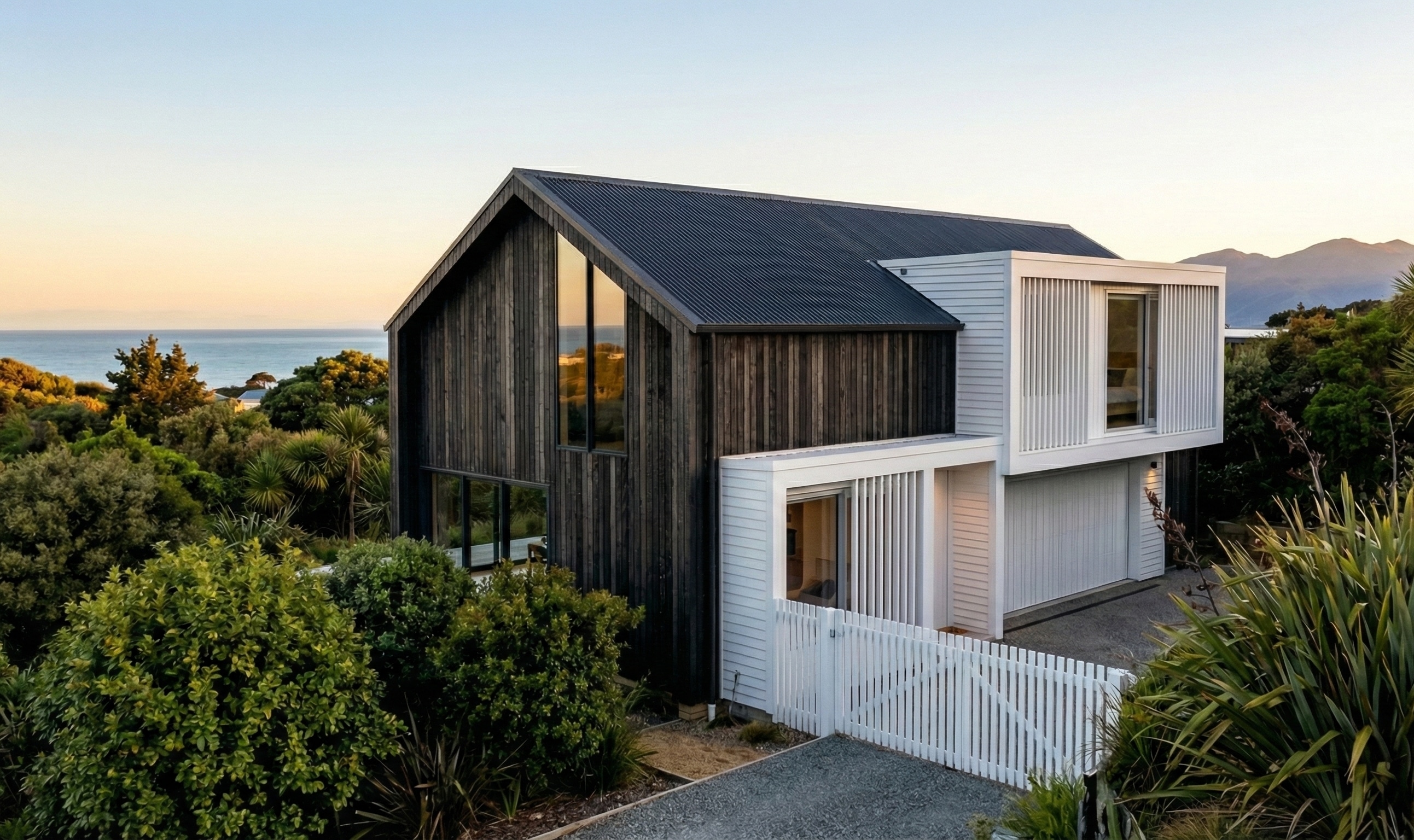 Two-storey coastal house with dark timber gable volume and white box-like extension, surrounded by lush greenery, overlooking the ocean at sunset with a white fence and gravel driveway in the foreground